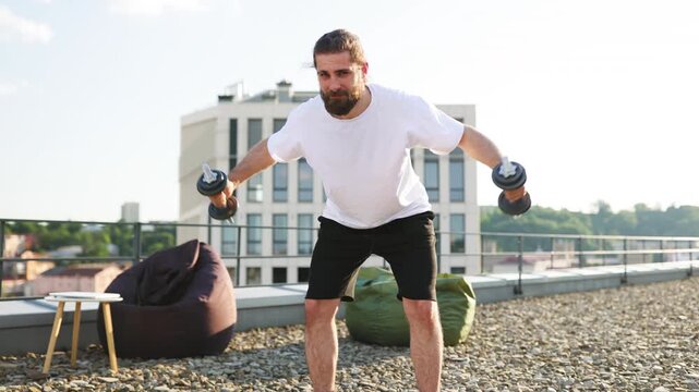 A bearded man with his hair tied back is performing a lateral raise exercise with dumbbells on a rooftop terrace with a city view