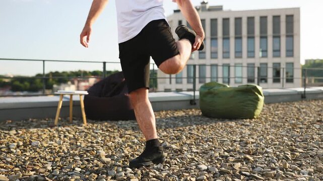 A man in athletic wear performs a quad stretch on a gravel-covered rooftop with a modern building in the background