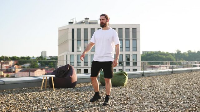 A man with a beard performs a side stretch on a gravel rooftop with beanbag chairs and a modern building in the background