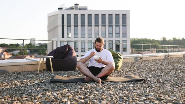 A man with a beard sits cross-legged on a yoga mat on a gravel rooftop, eating a salad from a bowl while beanbag chairs and a modern building are visible in the background