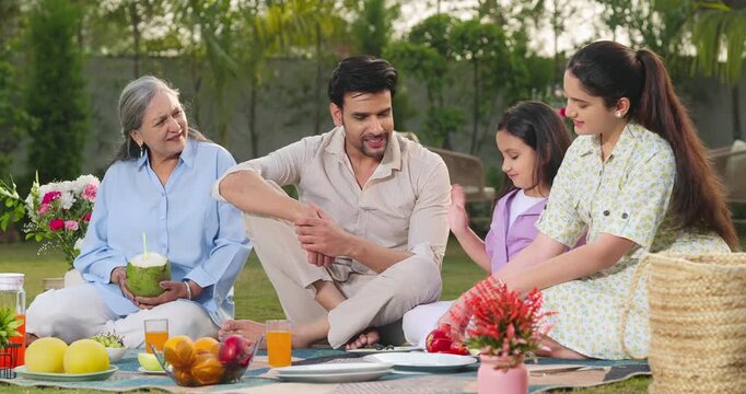 Indian man woman male group sitting on mat garden place talk speak enjoy trip have fun joy day old aged female drink coconut water young mom dad hold apple girl child kid eat fresh fruit spend time