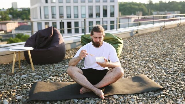 A man with a beard and bun sits cross-legged on a yoga mat on a gravel rooftop, eating a salad from a bowl while beanbag chairs and a modern building are visible in the background