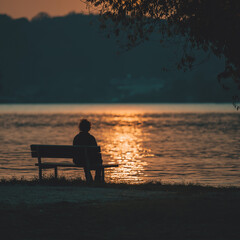 Obraz premium Solitude at Sunset: A lone individual finds solace on a bench overlooking the tranquil expanse of water at dusk, immersed in the serene ambiance of the twilight.