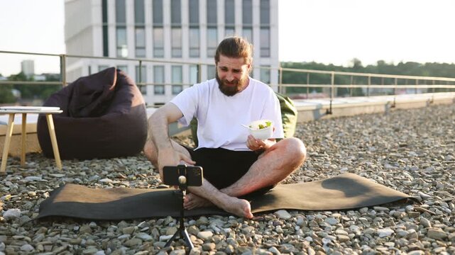 A man with a beard sits cross-legged on a yoga mat on a gravel rooftop, holding a container of food and talking to a smartphone on a tripod
