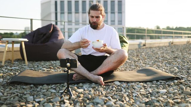 A man with a beard and bun sits cross-legged on a yoga mat on a gravel rooftop, eating a salad from a bowl while filming himself on a smartphone
