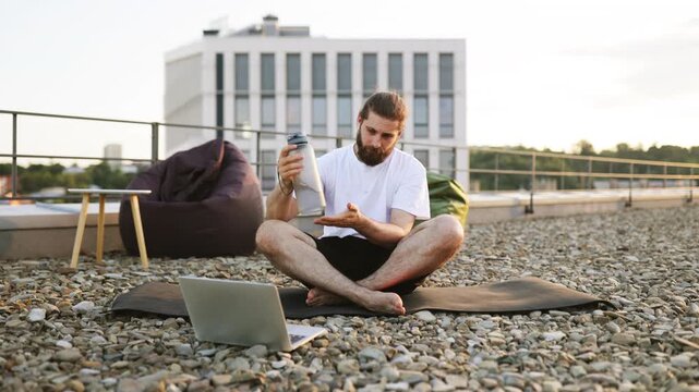 A bearded man with his hair in a bun sits cross-legged on a yoga mat on a gravel rooftop, holding a water bottle and looking at his laptop