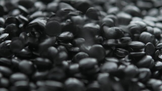 Black plastic pellets pour from above onto resin granules pile in macro view, showing raw polymer material texture closeup.
