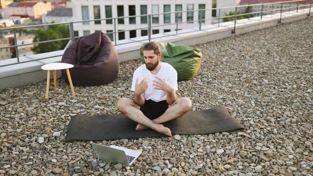 A man with a beard and bun hairstyle sits cross-legged on a yoga mat on a gravel rooftop, engaging in a video call on his laptop with beanbag chairs nearby
