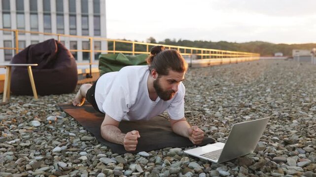 A man with a beard and bun hairstyle performs a plank exercise on a yoga mat on a rooftop while looking at a laptop screen