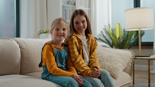 Portrait two happy Caucasian sisters siblings smiling joyful relaxing on couch looking at camera at home cheerful children little girls daughters kiss smile posing in living room together good family