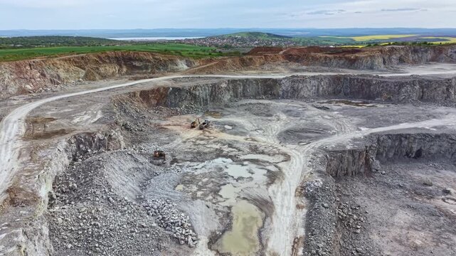 Heavy machinery works in a rock quarry. Large areas of land show signs of excavation. The landscape includes rocks, soil, and a pool of water under a cloudy sky.