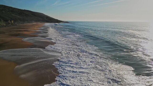 Slow motion drone footage of Praia do Salgado beach in Nazar&eacute;, Portugal. Cinematic waves crashing on the Atlantic coast with beautiful morning light over the rugged landscape