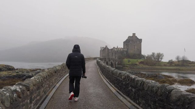 Person walking along stone path toward Eilean Donan Castle in misty Scottish landscape