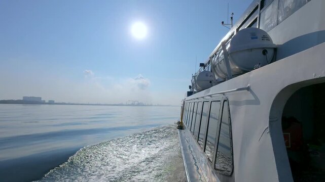 Point-of-view shot from a moving ferry boat deck showing a crisp white foam wake and a distant urban skyline along a major shipping lane in the Singapore Strait.