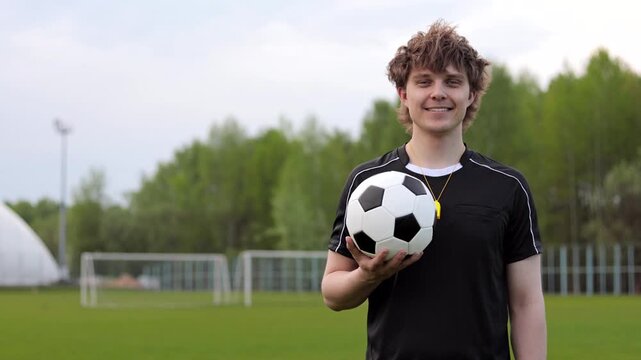 Smiling young man referee or coach in black football uniform holding a soccer ball on a field. Copy space.