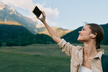Obraz premium A woman takes a confident selfie in a bright mountain meadow, wearing a beige jacket and backpack, headphones on, enjoying the fresh air and expansive scenery.