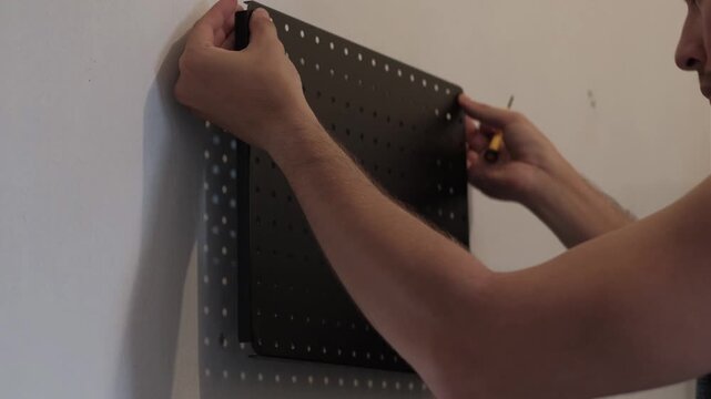 Medium shot of hands installing black pegboard on wall using screwdriver indoors for home improvement and workspace organization.