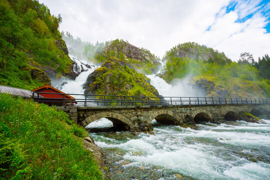 Powerful Twin Waterfall Crashing Over Rocky Cliff with Stone Bridge and Red Cabin in Norway
