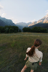 Obraz premium Young woman with long brown hair walks through a grassy field surrounded by mountains during a clear day, enjoying a peaceful outdoor lifestyle moment.