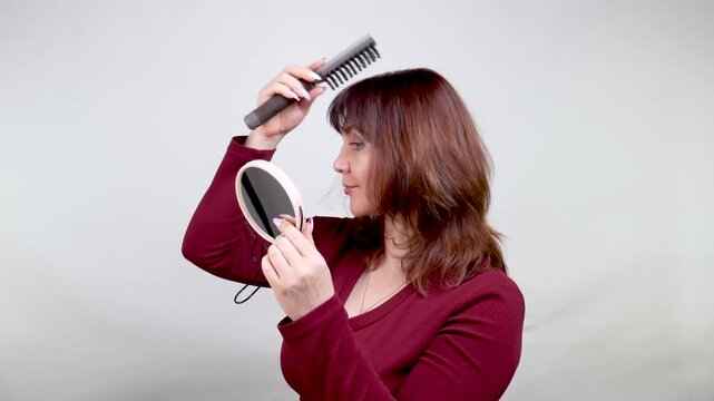 A woman in her 40s or 50s with a comb on a neutral background combing her hair in front of a mirror