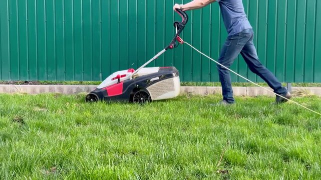 Man pushing an electric lawn mower along a green metal fence in the garden