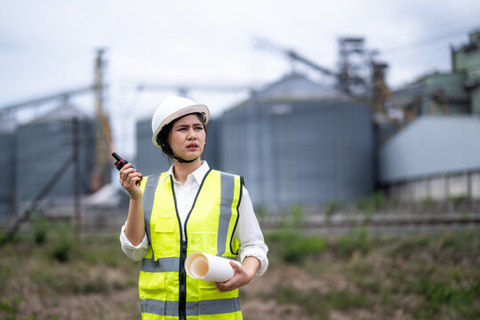 Female Engineer Wearing Safety Helmet and Vest Holding Documents and Walkie Talkie at Construction Site