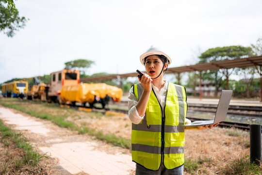 Female railway worker with safety vest talking into walkie talkie and holding a laptop outdoors