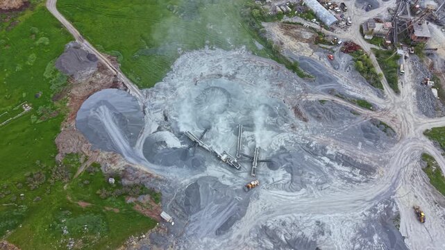 A construction site shows heavy machinery working on a gravel extraction project. Large areas of gray material are visible, with some equipment in operation under a clear sky.