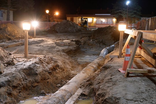 Night construction site with exposed pipe and dirt. Workers operate machinery nearby under artificial lights. Scene shows active excavation and groundwork