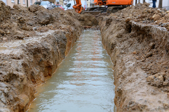 Mud-filled trench runs straight through dirt. Water flows slowly between earthy sides. Orange machinery rests nearby, idle. Workers stand at trench's edge, unseen