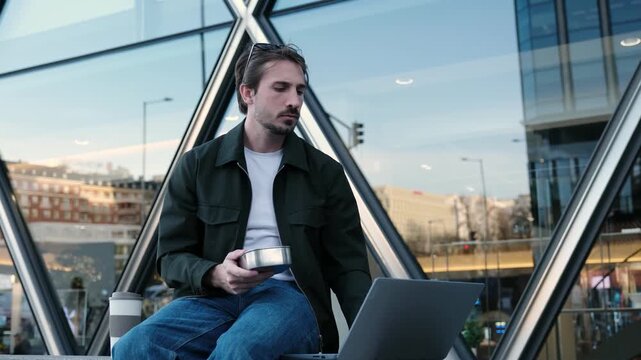 Young man having a meal from a reusable container while working on his computer in the city