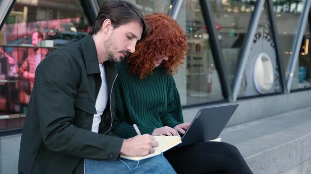 Happy students working together on a project, sitting on city steps with a computer and notepad