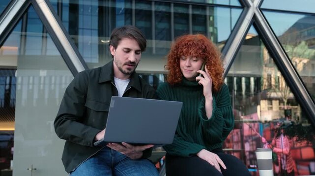 Two young professionals working together on a laptop with a modern glass building in the background