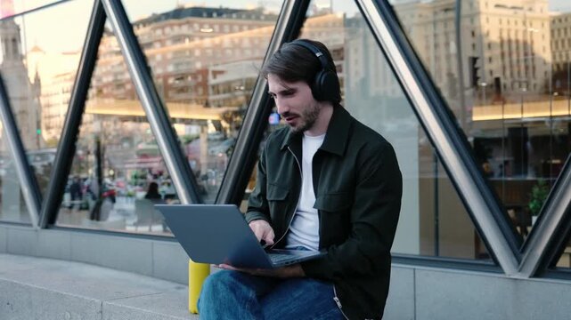 Male freelancer listening to music on headphones while typing on a laptop in a modern city street