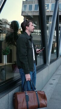 Young man waiting on a city street while checking his mobile phone and holding a brown leather bag