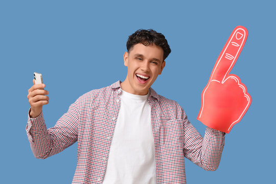 Happy young man with foam finger and mobile phone pointing at something on blue background. Sports bet concept