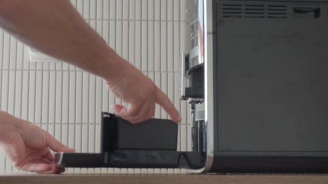 Hands removing black container from side compartment of coffee machine on kitchen counter. Close-up of household appliance cleaning maintenance and daily coffee preparation.