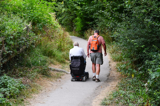 Wheel chair walk, man on mobility scooter out enjoying the English countryside with his able bodied friend as they walk and chat together on a summers day.