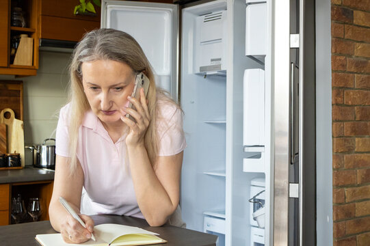 Woman talks to repair technician on phone and writes appointment details in notebook. Appliance service scheduling, household problem solving and maintenance planning concept