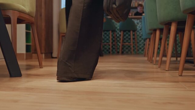 Closeup wooden floor walking businessperson shoes under chairs, soft ambient lighting, midcentury chairs and tables, polished wood planks, measured footsteps and steady pace, briefcase swing,