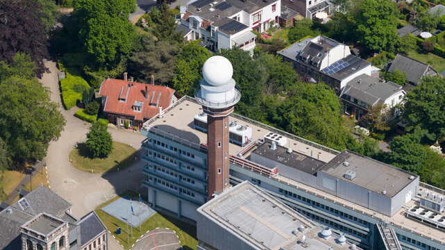 Aerial view of the KNMI building featuring its iconic radar tower and white dome surrounded by lush green trees and residential houses in De Bilt, Utrecht, Netherlands.