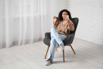 Obraz premium Young African-American woman with mobile phone sitting in grey armchair at home