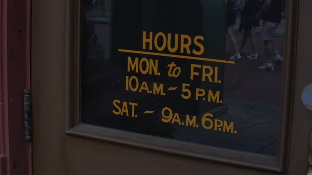 Hours sign displayed on a storefront door with business schedule in gold lettering. Reflections of pedestrians walking past appear in the dark glass window