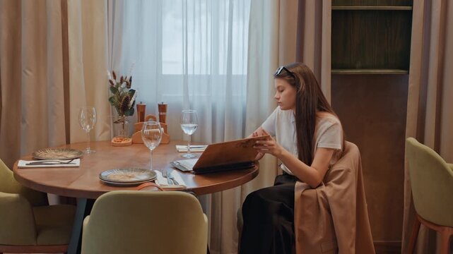 Young woman at dining table unpacking wooden box, soft natural light, set plates and glasses, beige coat draped over chair, window curtains, calm introspective expression, preparing for meal