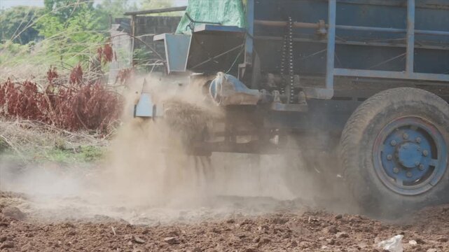 Side view of a blue agricultural fertilizer spreader machine discharging organic manure or fertilizer onto a brown field in a rural farm setting under bright natural daylight outdoors.