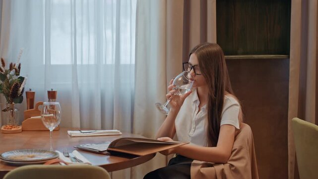 Caucasian woman sipping water at table with menu in lap, pauses between sips and scans pages relaxed and thoughtful moment suggesting student, business traveler, or critic in modern interior