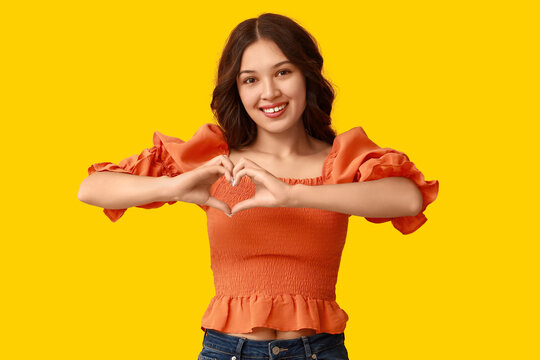 Happy young Asian woman showing heart gesture on yellow background. Valentine's Day celebration