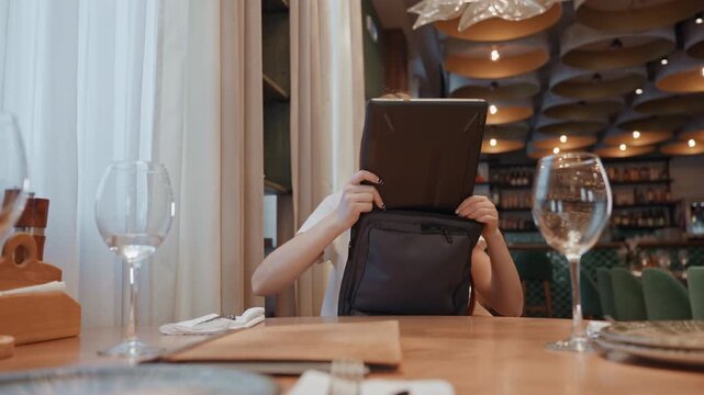 Young woman checking laptop privacy in restaurant, lifting device from bag and scanning surroundings, wine glasses and place setting in foreground, ambient ceiling lights and modern interior, cautious