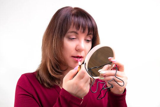 A brunette woman with a mirror applies pink lipstick to her lips while doing her makeup