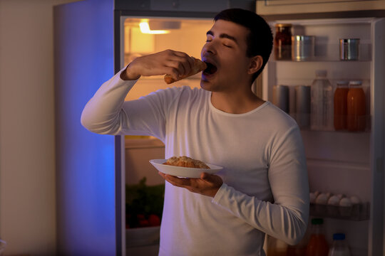 Hungry young man eating croissant near open fridge in kitchen at night